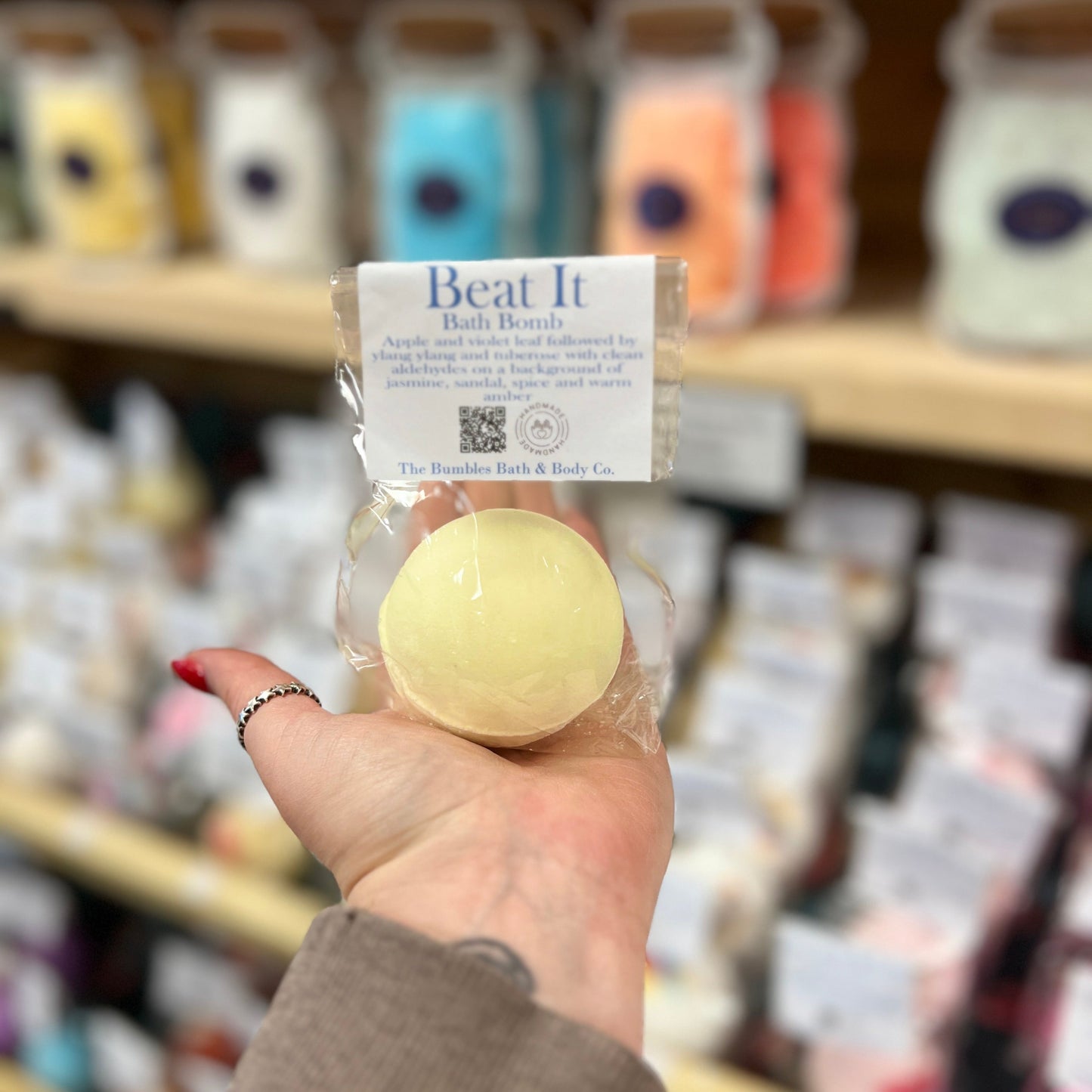 Hand holding a yellow bath bomb labeled 'Beat It' in front of shelves with various products.