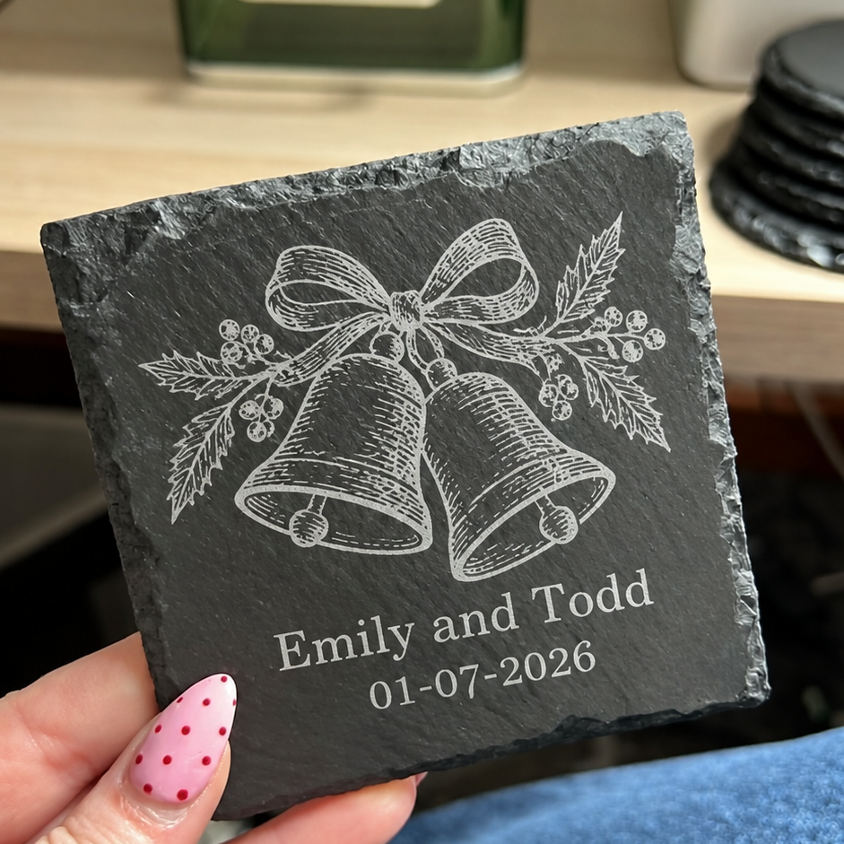 A hand holding a square dark slate coaster engraved with two hanging bells, a bow, holly leaves, and the personalized text 'Emily and Todd 01-07-2026'. The background shows a blurred desk with a green bottle and blue jeans.