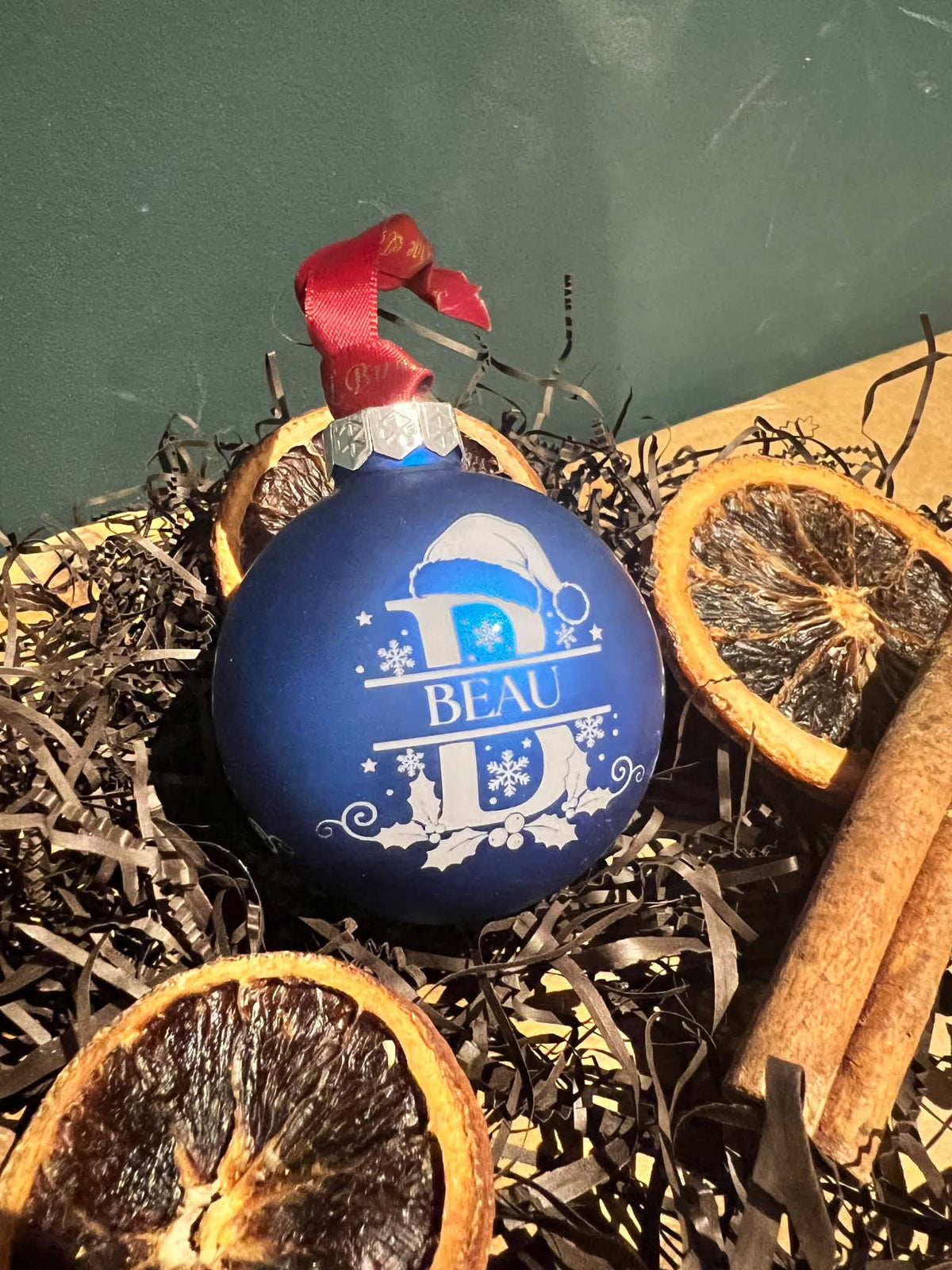 A blue Christmas ornament with a white monogram design reading 'BEAU' and a red ribbon hanger, styled in shredded black paper with dried orange slices and cinnamon sticks.