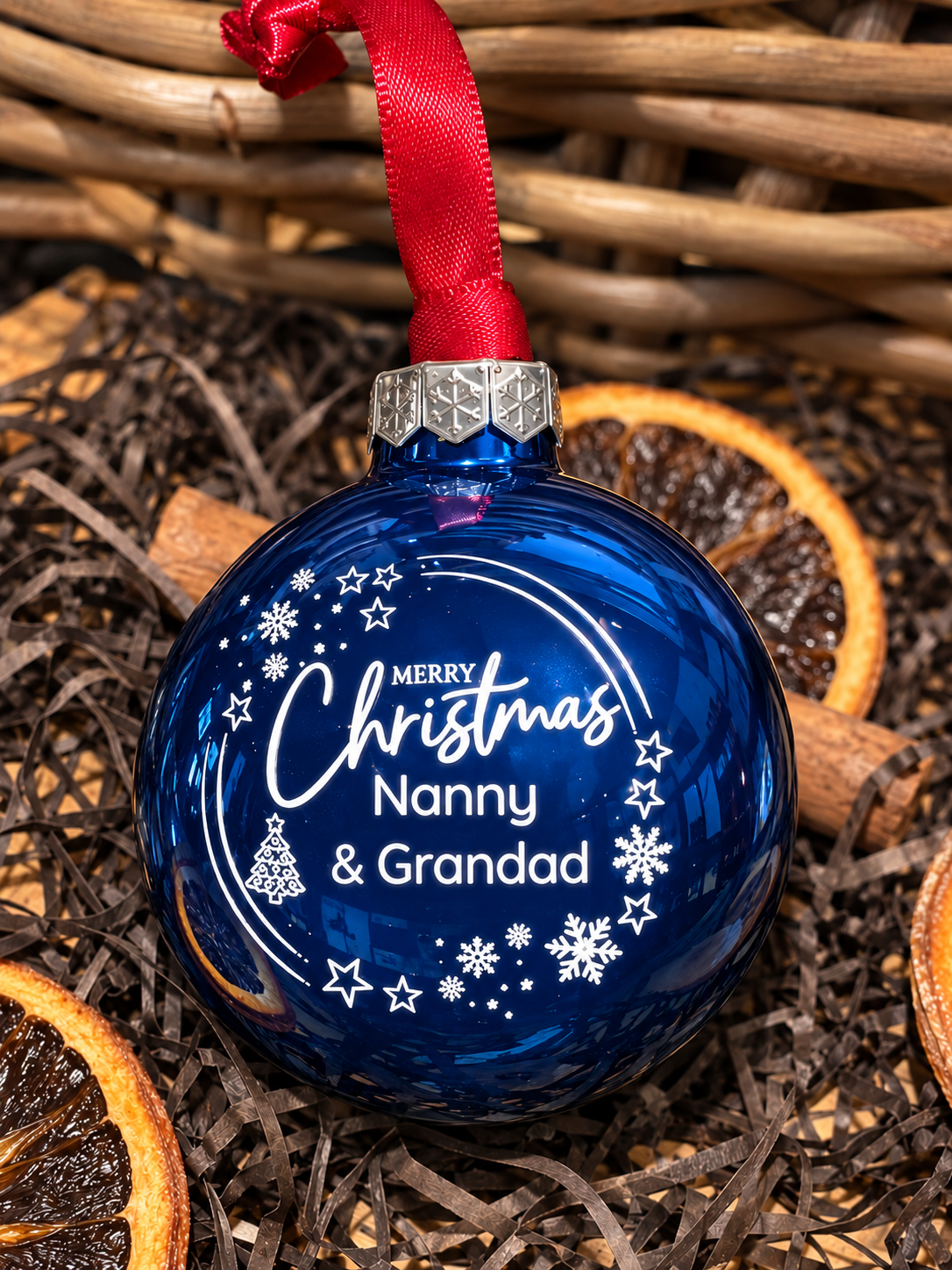 A personalized blue Christmas ornament with a red ribbon hanger, featuring white text that reads 'Merry Christmas Nanny & Grandad' with snowflake and star designs, styled on a bed of dried orange slices and twine.