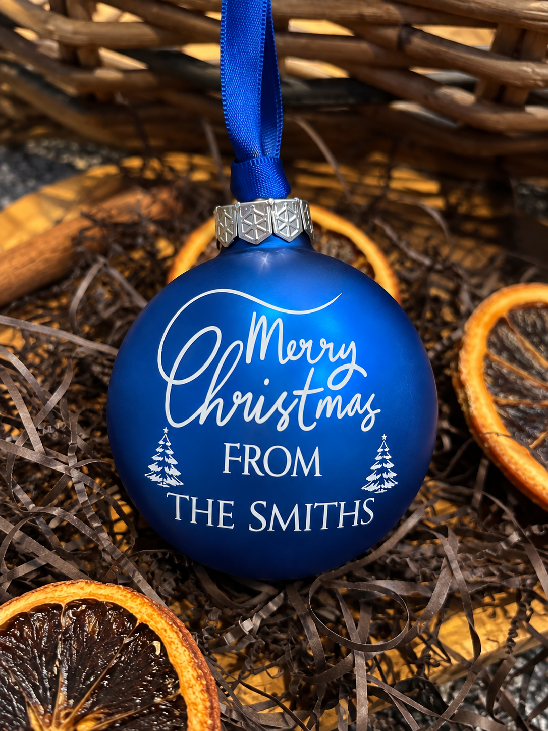A personalized royal blue Christmas ornament with a blue ribbon hanger, featuring the text 'Merry Christmas FROM THE SMITHS' in white script and block lettering, decorated with small white Christmas tree illustrations. The ornament is styled in a flat-lay arrangement with dried orange slices and brown shredded paper.