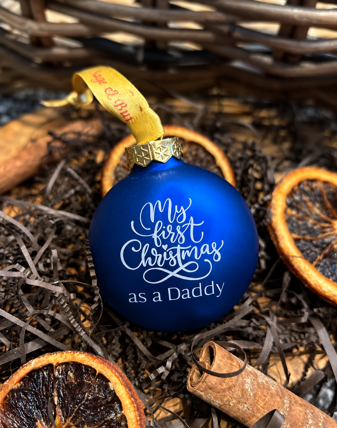 A blue Christmas ornament with white script text reading 'My first Christmas as a Daddy' and a yellow hanging ribbon, styled among dried orange slices, cinnamon sticks, and shredded brown filler.