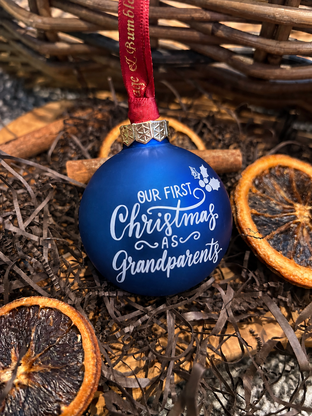 A round blue Christmas ornament with white script text reading 'Our First Christmas As Grandparents' hanging from a red ribbon, styled in dried orange slices and brown paper shreds.