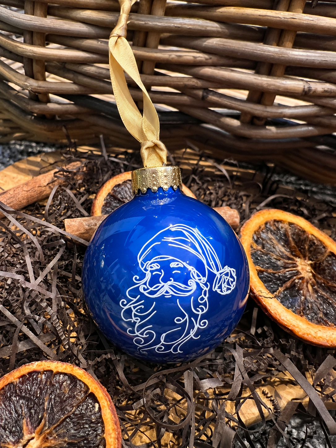 A glossy cobalt blue Christmas ornament featuring a white line-art illustration of Santa Claus, hanging from a beige ribbon. The ornament is styled in a rustic setting with dried orange slices, cinnamon sticks, and shredded paper in front of a wicker basket.
