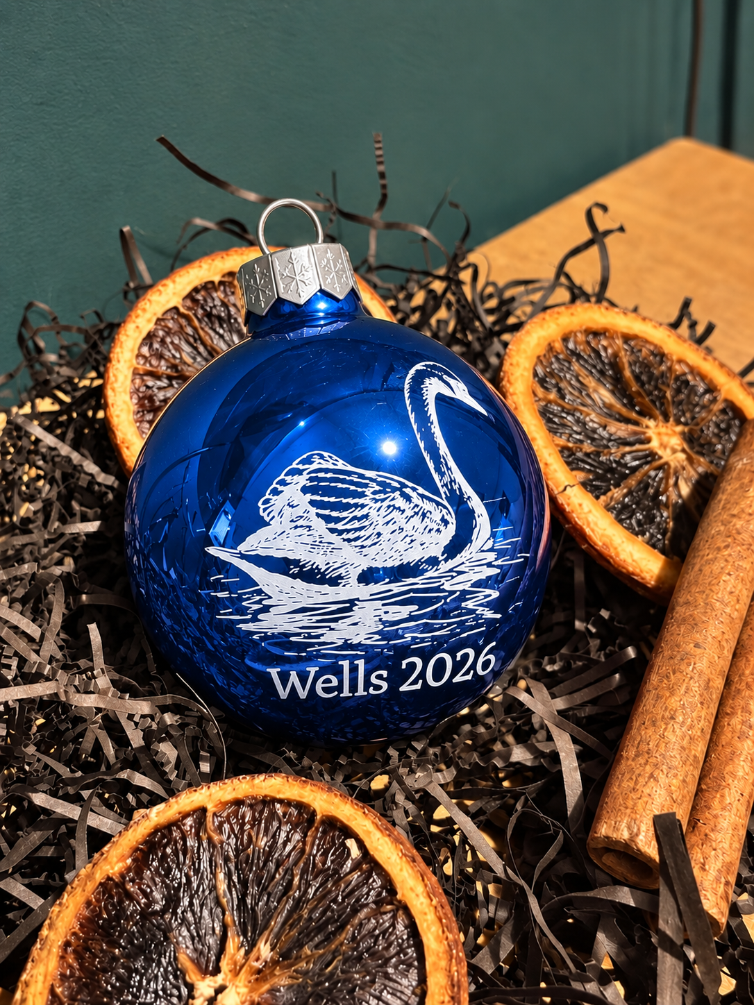A personalized royal blue glass Christmas ornament featuring a white engraved swan and the text 'Wells 2026', styled in a holiday setting with dried orange slices, cinnamon sticks, and black shreds on a wooden surface.