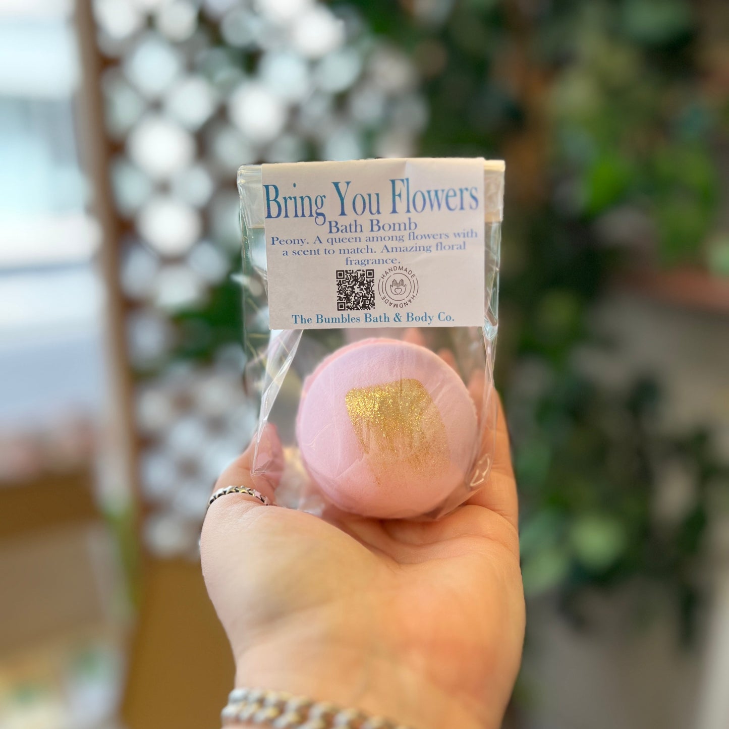 A hand holding a packaged pink bath bomb labeled 'Bring You Flowers Bath Bomb' with a gold foil flower design, against a blurred outdoor background with greenery.