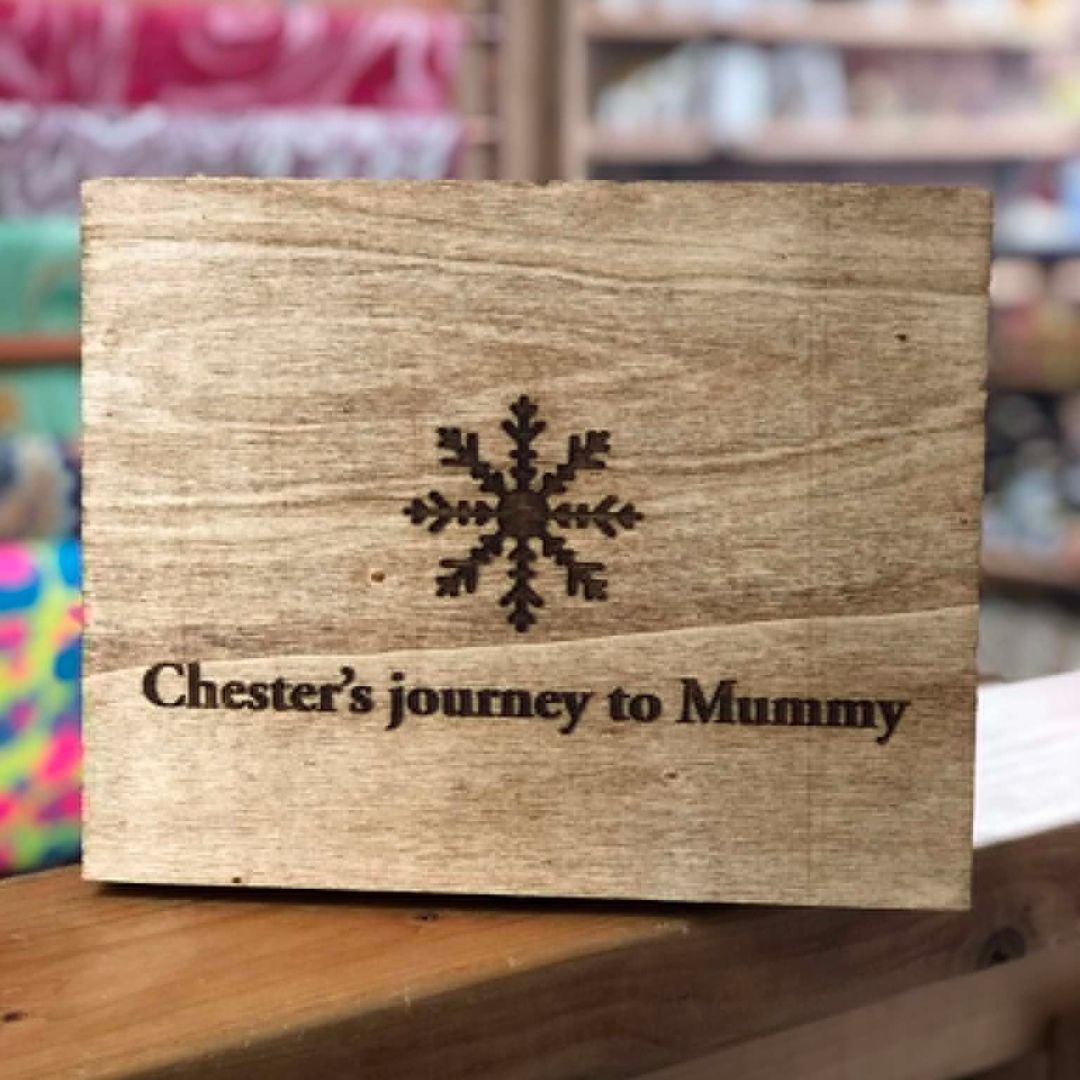 A rectangular wooden keepsake box lid engraved with a snowflake and the text 'Chester's journey to Mummy', shown standing on a wooden surface with a blurred shop background.