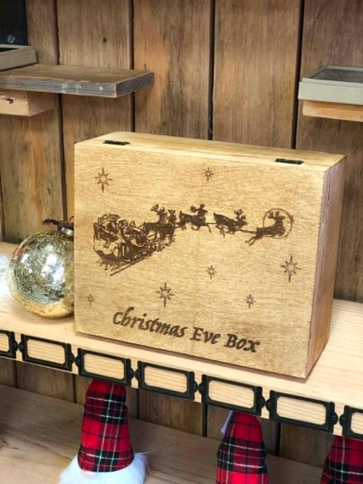A rectangular wooden Christmas Eve box with a laser-engraved Santa Claus and reindeer illustration, displayed on a wooden shelf next to a metallic ornament.