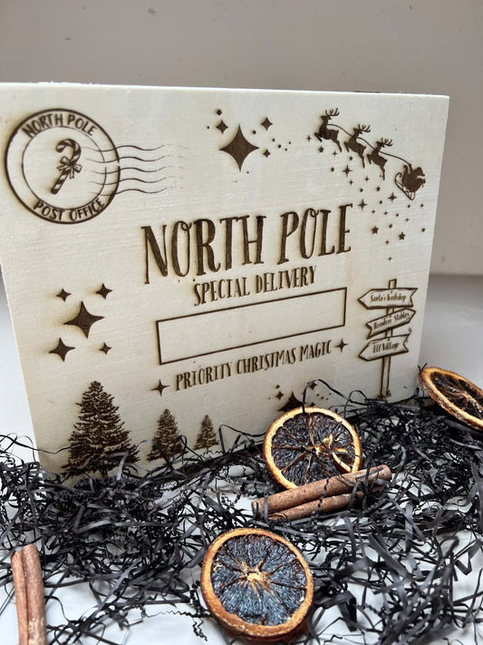A rectangular cream-colored Christmas postcard or card insert with laser-engraved holiday illustrations and the text 'NORTH POLE SPECIAL DELIVERY' and 'PRIORITY CHRISTMAS MESSAGE,' styled on black tinsel with dried orange slices and cinnamon sticks.