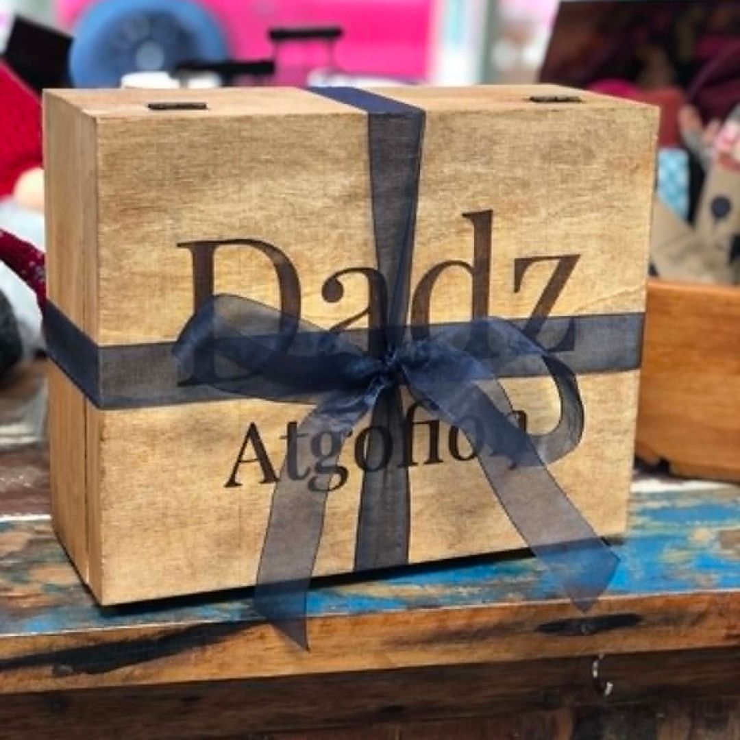 A wooden gift box with the words 'Dadz Atgofion' printed on the front, wrapped with a sheer dark gray ribbon, shown on a rustic wooden table.