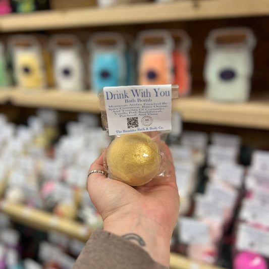 Hand holding a gold bath bomb labeled 'Drink With You' in front of a store shelf with various products.