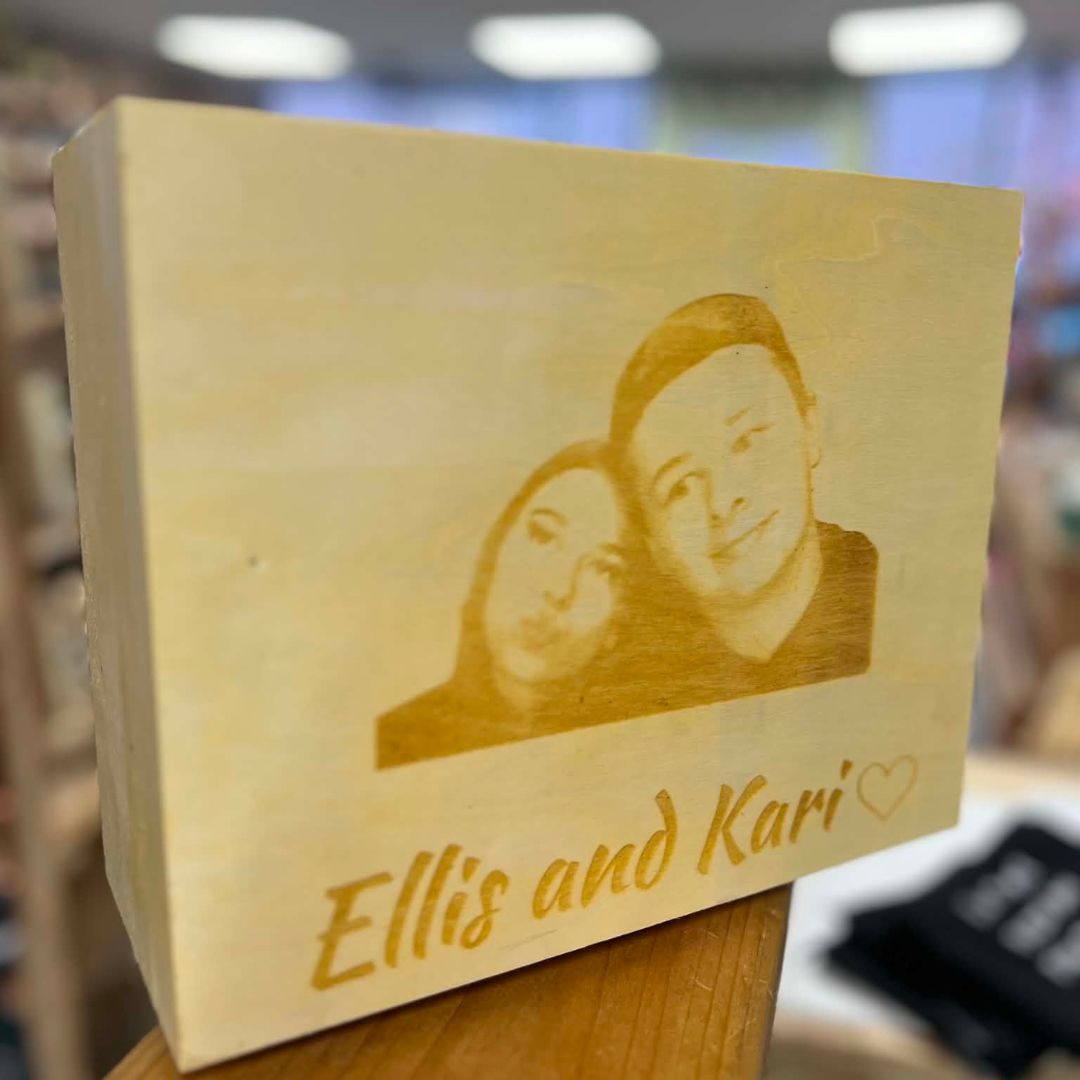 A rectangular wooden keepsake box with a custom laser-engraved portrait of a couple and the names 'Ellis and Kari' with a heart, displayed on a wooden surface with a blurred store background.