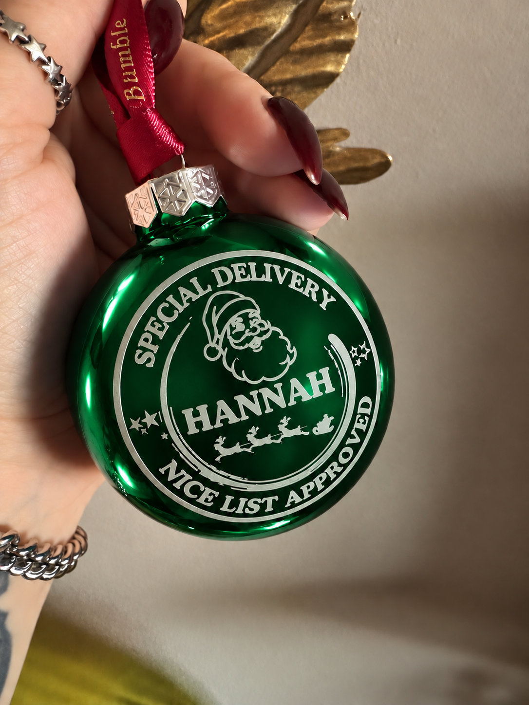 A hand holds a personalized green Christmas ornament with a red hanging ribbon. The ornament features a white circular print with Santa's face, sleigh, and the text 'SPECIAL DELIVERY HANNAH NICE LIST APPROVED'.