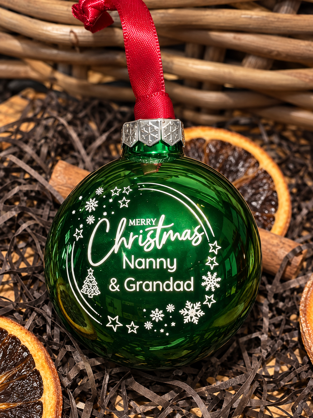 A personalized green glass Christmas ornament with a red ribbon hanger, featuring white printed text that reads 'Merry Christmas Nanny & Grandad' surrounded by stars and snowflakes. The ornament is styled in a holiday setting on a bed of brown shredded paper and dried orange slices, with a wicker basket in the background.