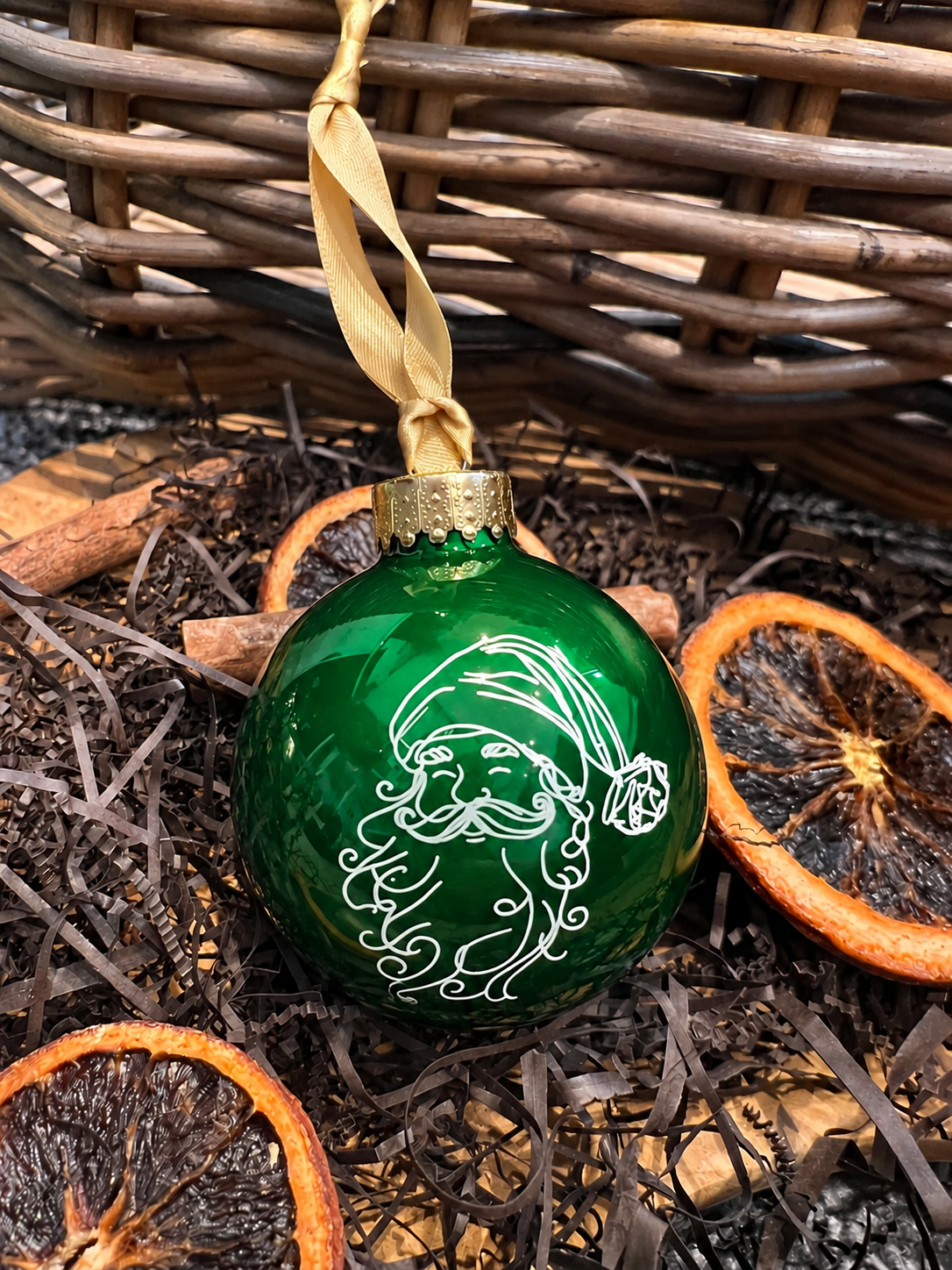 A green Christmas ornament shaped like a Santa Claus face, hanging from a gold ribbon in front of a wicker basket with dried orange slices and cinnamon sticks.