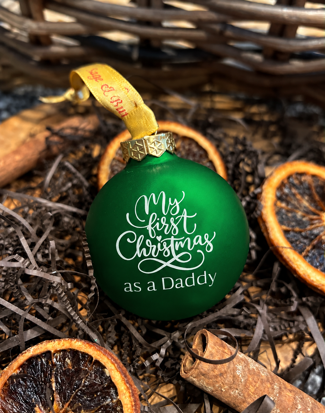 A green Christmas ornament with white script text reading 'My first Christmas as a Daddy' and a gold cap, resting in dried orange slices and cinnamon sticks.