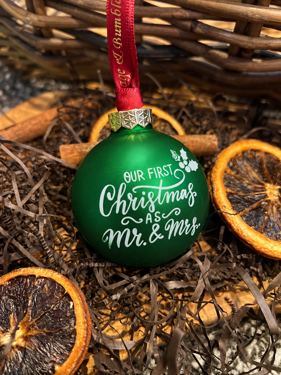 A green Christmas ornament with a red ribbon, featuring the white printed text 'OUR FIRST Christmas AS Mr. & Mrs.