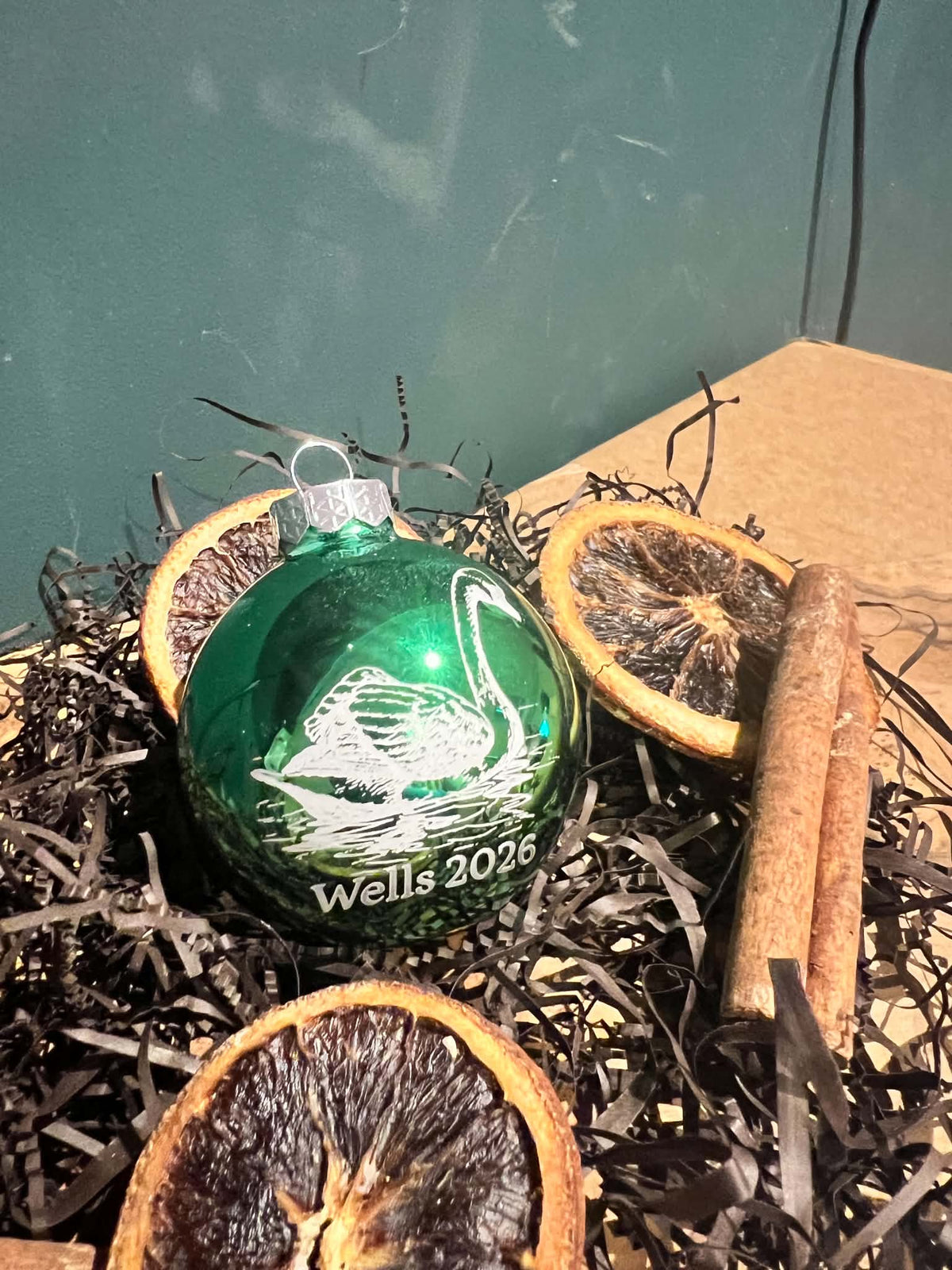 A green glass Christmas ornament with a white swan design and the text 'Wells 2026' resting in dark shredded paper with dried orange slices and cinnamon sticks.