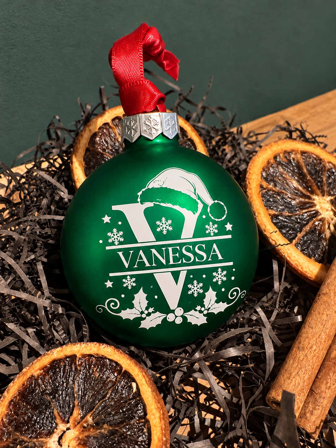A personalized Christmas ornament shaped like a green bauble with a red ribbon loop, featuring a white monogram 'V' and the name 'VANESSA' with holiday motifs. The ornament is styled in dark shreds with dried orange slices and cinnamon sticks against a dark green background.