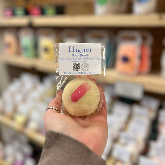 Hand holding a bath bomb labeled 'Higher' with a blurred store shelf in the background