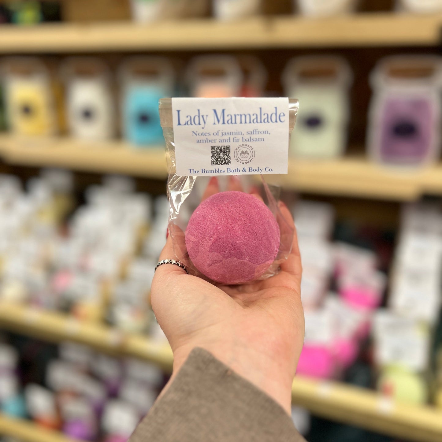 Hand holding a pink bath bomb labeled 'Lady Marmalade' in front of a store shelf.