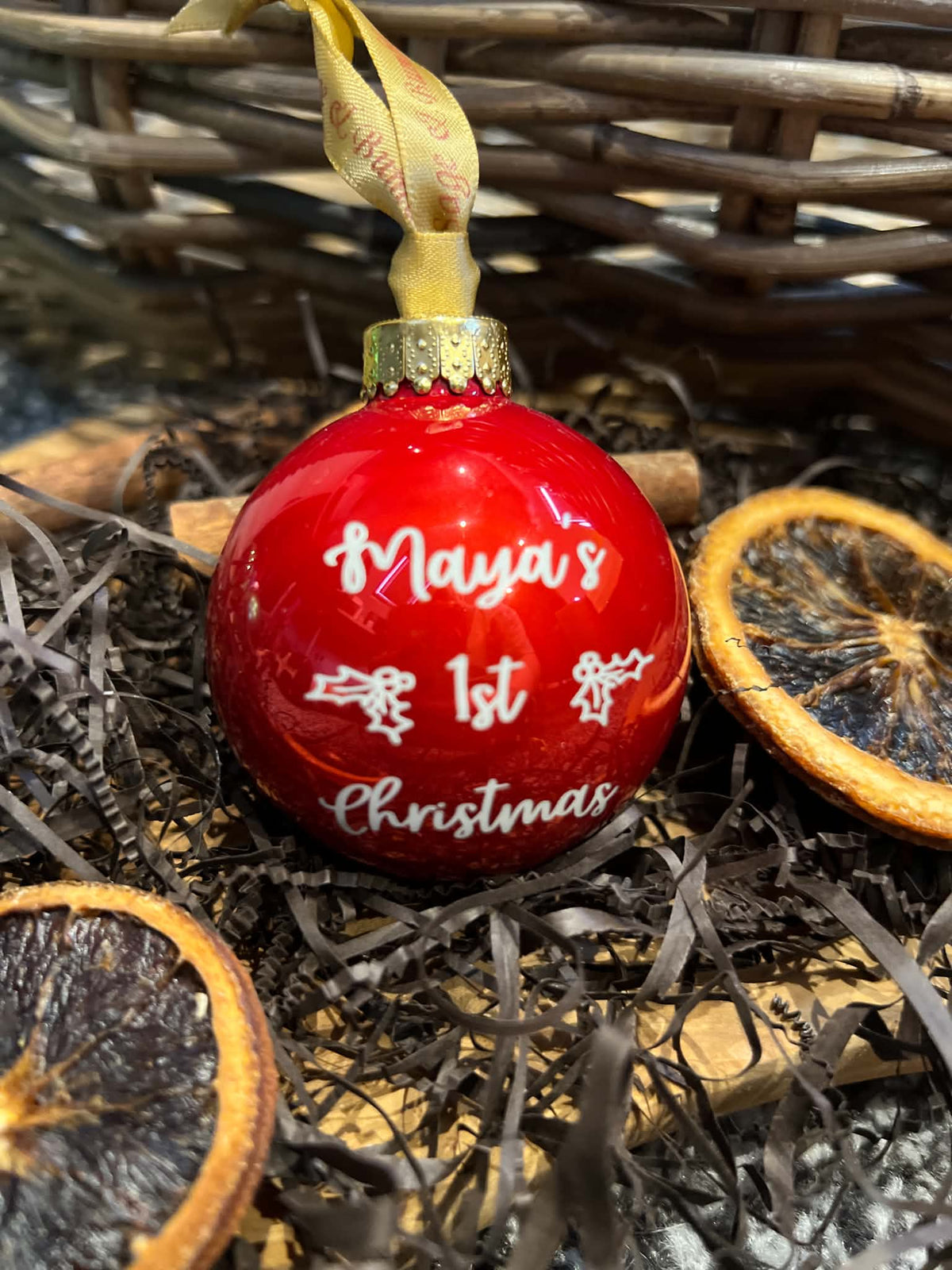 A glossy red Christmas ornament with white cursive text reading 'Maya's 1st Christmas' and a gold hanging loop, styled in dark shredded paper with dried orange slices and a wicker basket in the background.