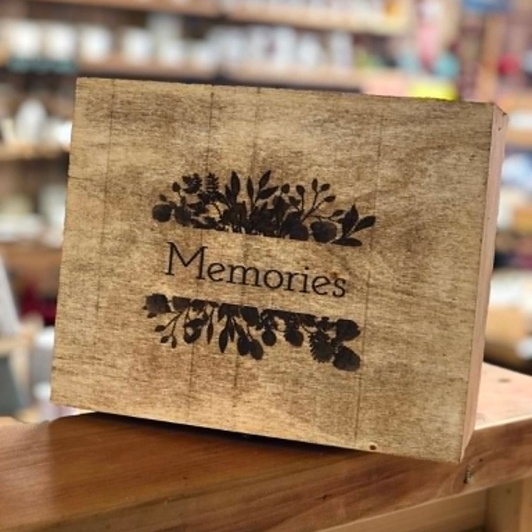 A rectangular rustic wooden keepsake box or plaque with the word 'Memories' engraved in the center, flanked by dark floral scroll designs. The box is shown standing on a wooden surface with a blurred store or shelf background.