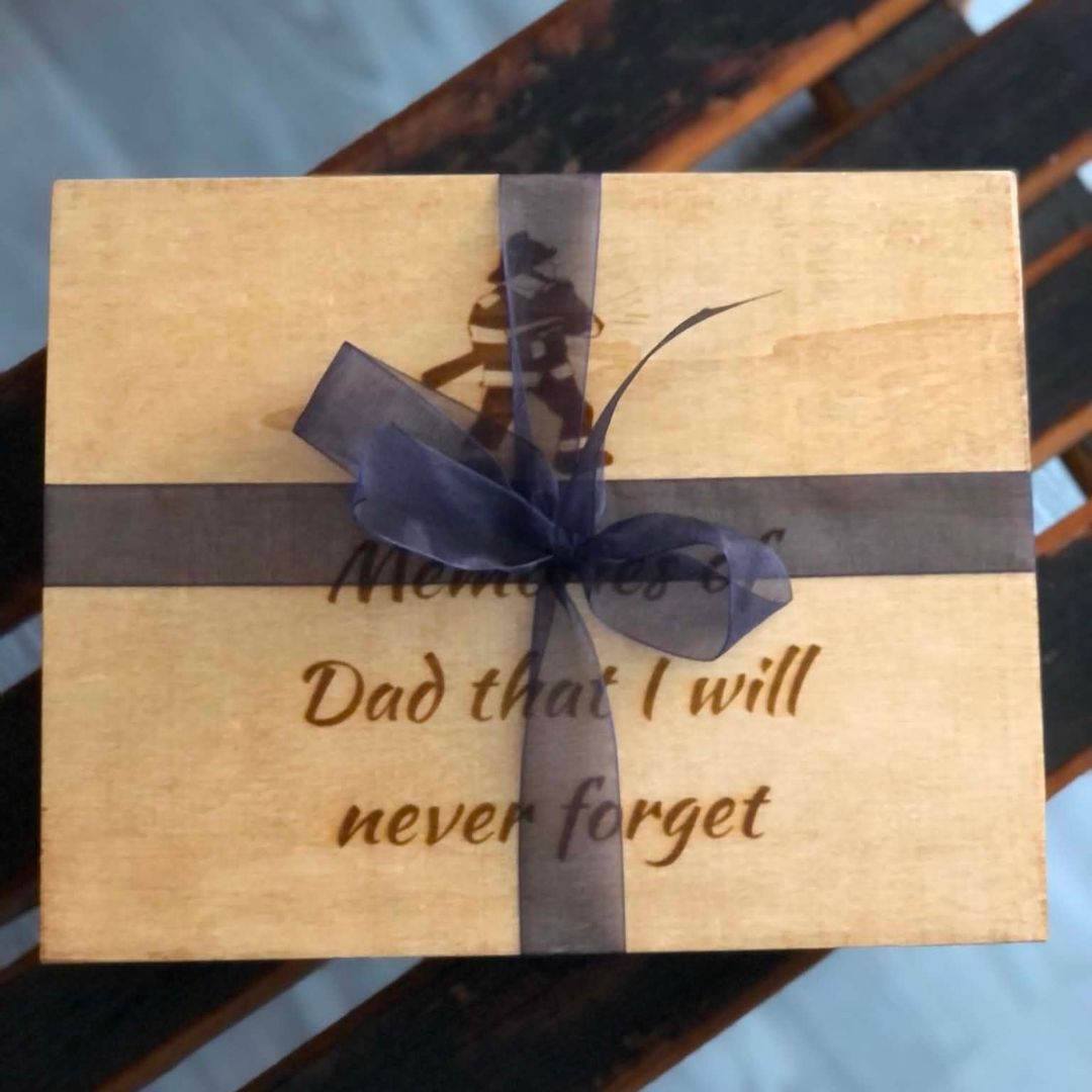 A square wooden keepsake box with engraved text reading 'Memories of Dad that I will never forget,' tied with a dark gray ribbon and bow, shown on a dark wooden slatted surface.