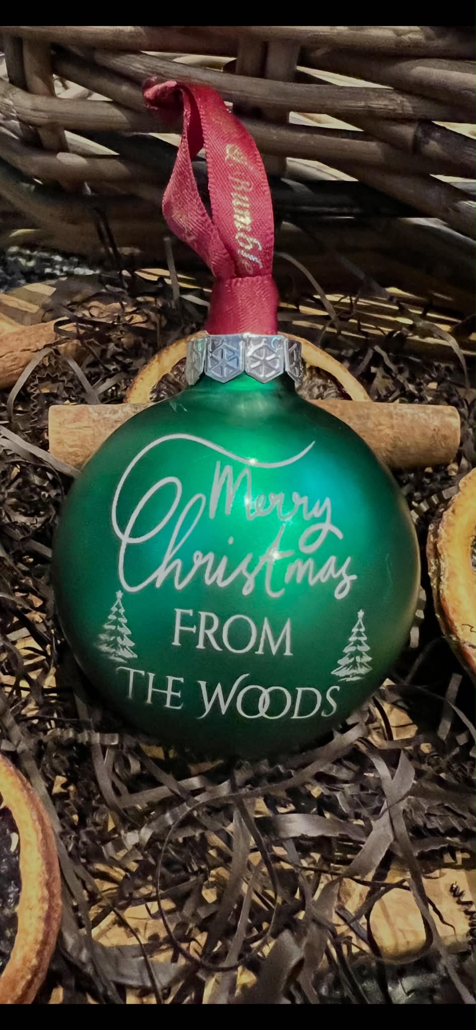A green Christmas ornament with a red ribbon hanger, featuring the text 'Merry Christmas FROM THE WOODS' in white script and font, displayed in a rustic setting with dried leaves and a wicker basket.