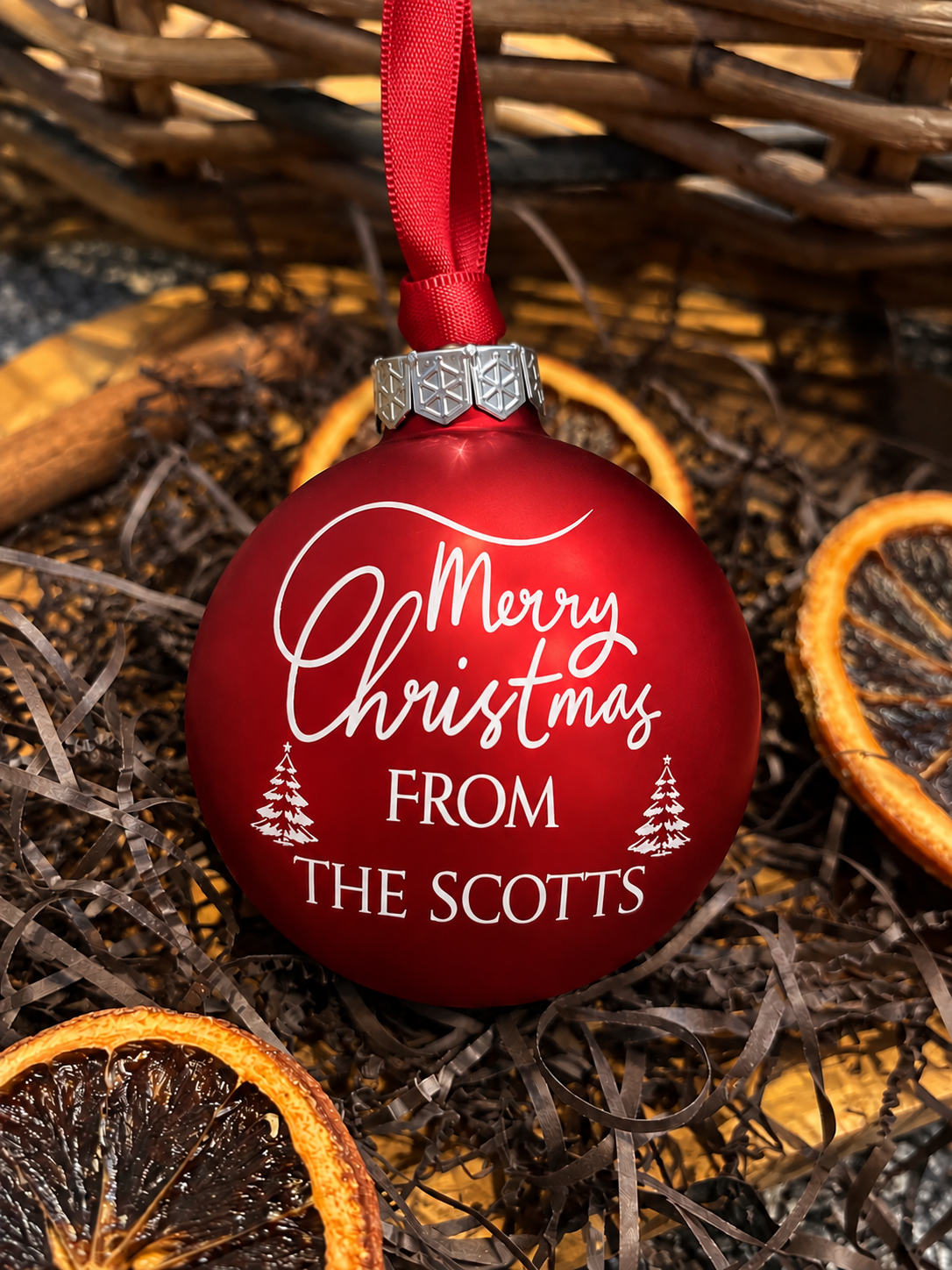 A red Christmas ornament with a red ribbon hanger, featuring the text 'Merry Christmas FROM THE SCOTTS' in white script and tree illustrations. The ornament is styled in a flat-lay setting on a bed of dried orange slices and brown twine.