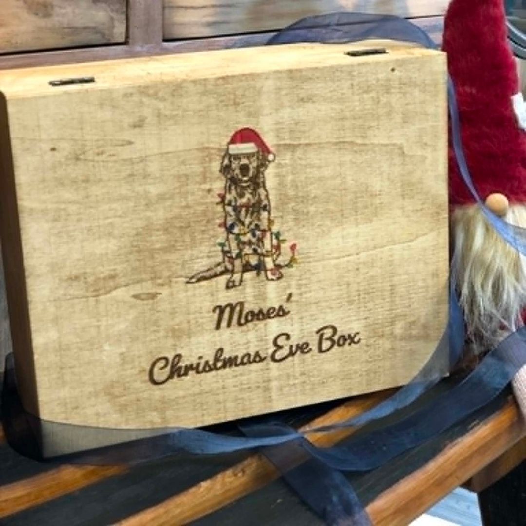 A wooden gift box engraved with a dog wearing a Santa hat and the words 'Moses' Christmas Eve Box,' tied with a dark ribbon and photographed on a wooden surface.
