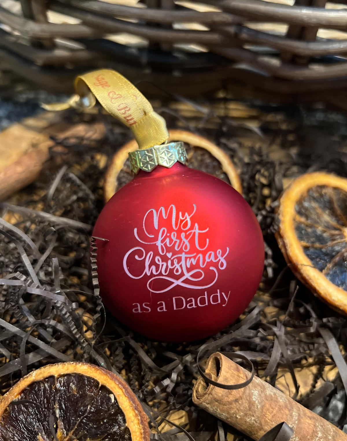 A red Christmas ornament with the text 'My first Christmas as a Daddy' in white script, resting on a holiday-themed background with dried orange slices, cinnamon sticks, and dark shreds.