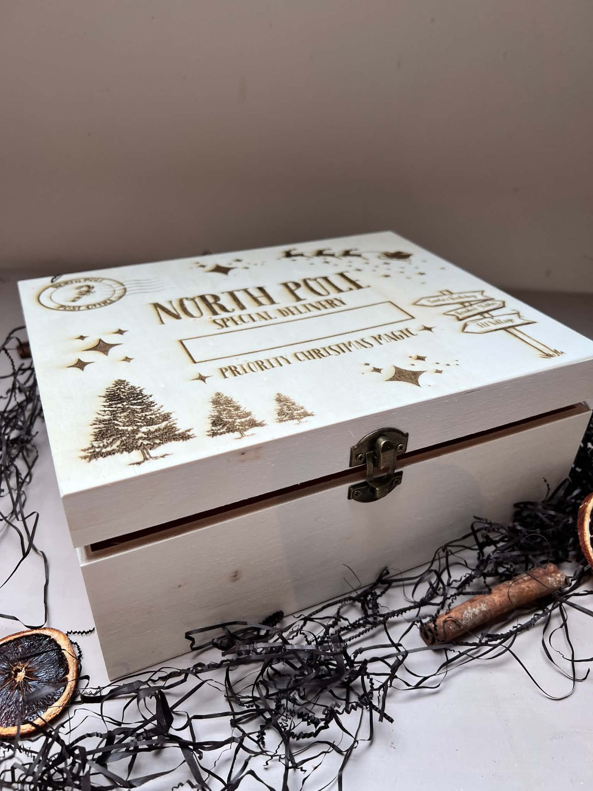 A white wooden keepsake box with a hinged lid, featuring laser-engraved Christmas illustrations and the text 'NORTH POLE SPECIAL ORDER' and 'Proudly Christmas 2021'. The box is styled on a white surface with black shredded paper, dried orange slices, and a cinnamon stick against a plain beige background.
