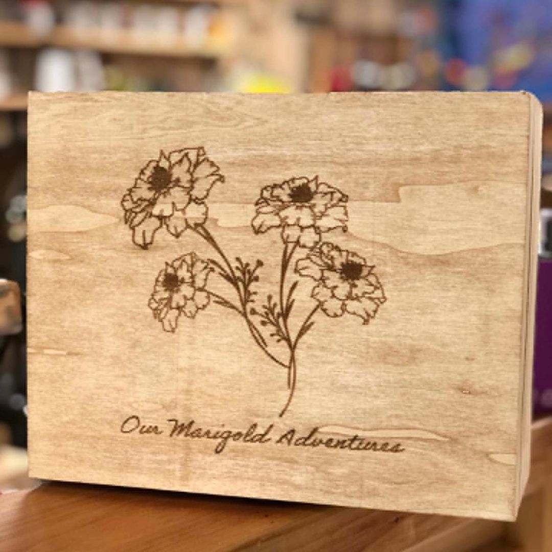 A square wooden keepsake box with a natural light wood finish, featuring a laser-engraved illustration of three marigold flowers and the text 'Our Marigold Adventures' on the front, set against a blurred background.