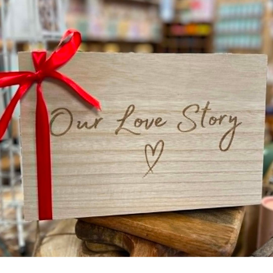 A rectangular light wood plaque engraved with the words 'Our Love Story' and a small heart, tied with a bright red ribbon bow, displayed on a wooden stand in a softly blurred shop setting.