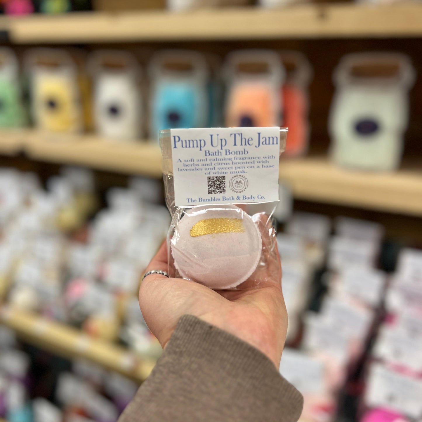 Hand holding a pink bath bomb labeled 'Pump Up The Jam' in a store setting.