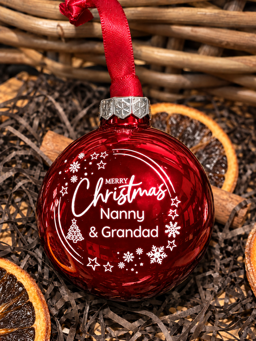 A red Christmas ornament with white personalized text reading 'Merry Christmas Nanny & Grandad' and white snowflake and star designs, hanging from a red ribbon. The ornament is styled on a bed of shredded brown paper with dried orange slices and a wicker basket in the background.