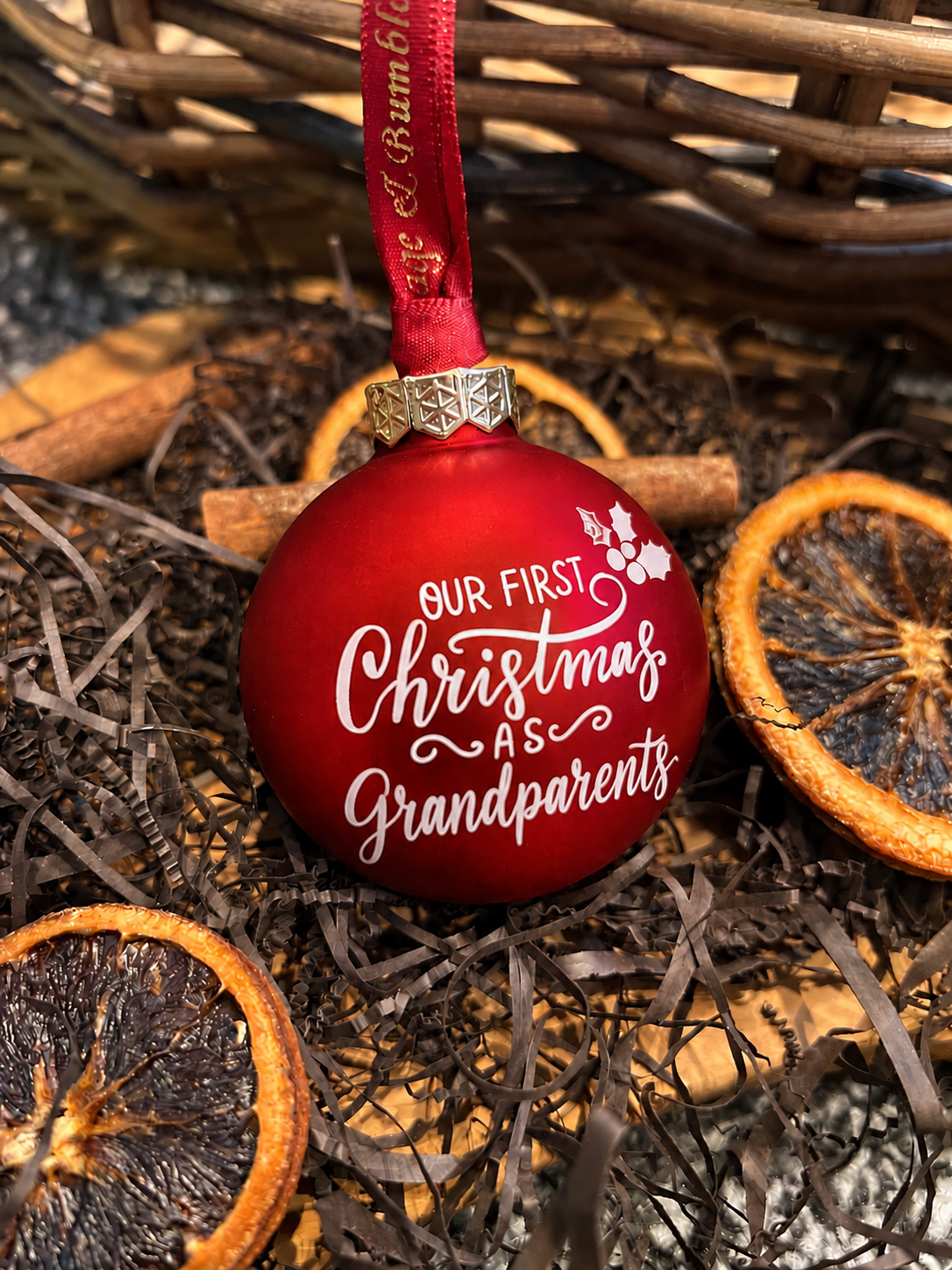 A red Christmas ornament with white script text reading 'OUR FIRST Christmas AS Grandparents' hanging from a red ribbon, styled in dried orange slices and brown paper shreds.