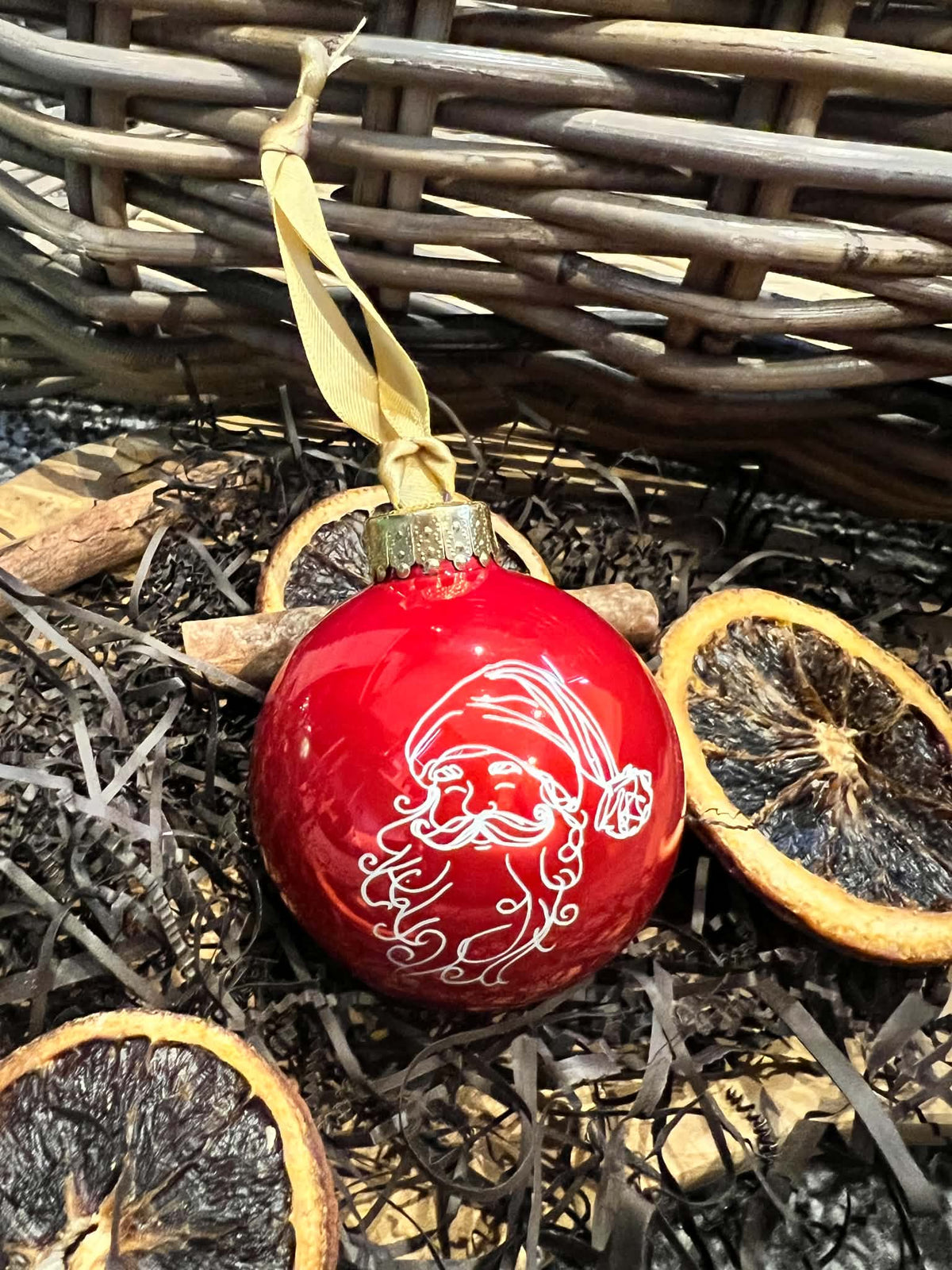 A glossy red Christmas ornament featuring a white hand-painted Santa Claus face, hanging from a beige ribbon in a rustic setting with dried orange slices and brown shredded paper.