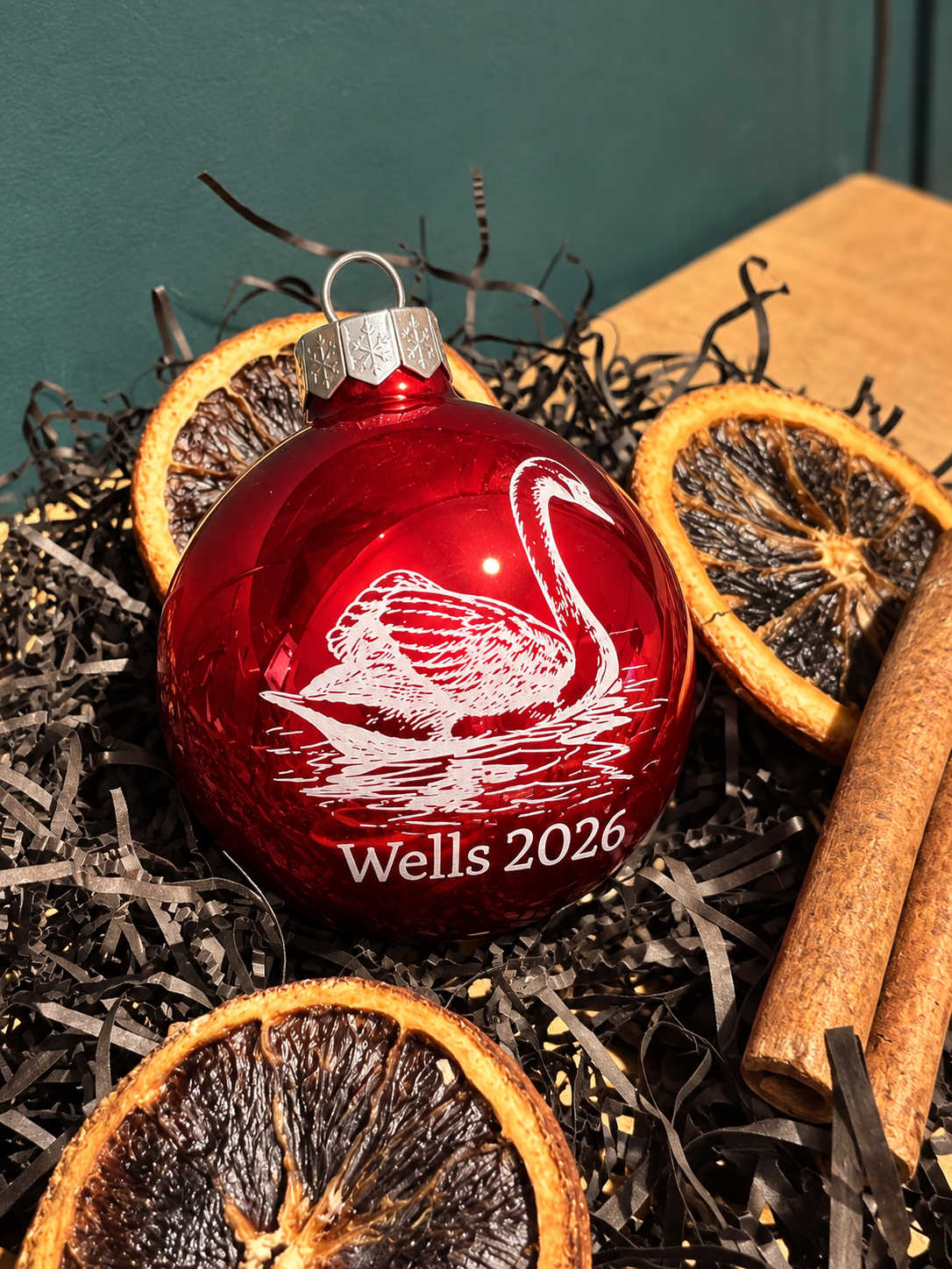 A red glass Christmas ornament with a white swan design and the text 'Wells 2026' resting on dark shredded paper with dried orange slices and cinnamon sticks.