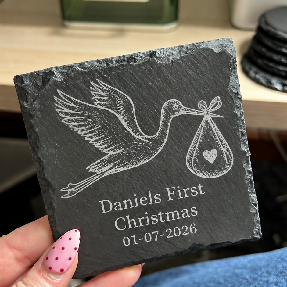 A hand with pink polka-dot nail polish holds a square dark slate coaster engraved with a stork carrying a baby bag and the personalized text 'Daniels First Christmas 01-07-2026'. The background shows a blurred desk environment with a green glass bottle and blue jeans.