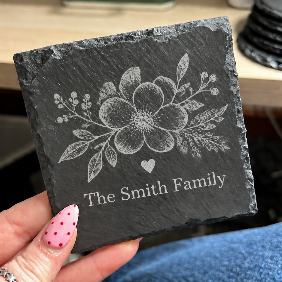 A hand with pink polka-dot nails holds a square dark slate coaster engraved with a floral design and the text 'The Smith Family'. The background shows a wooden desk with a green glass bottle and other household items.