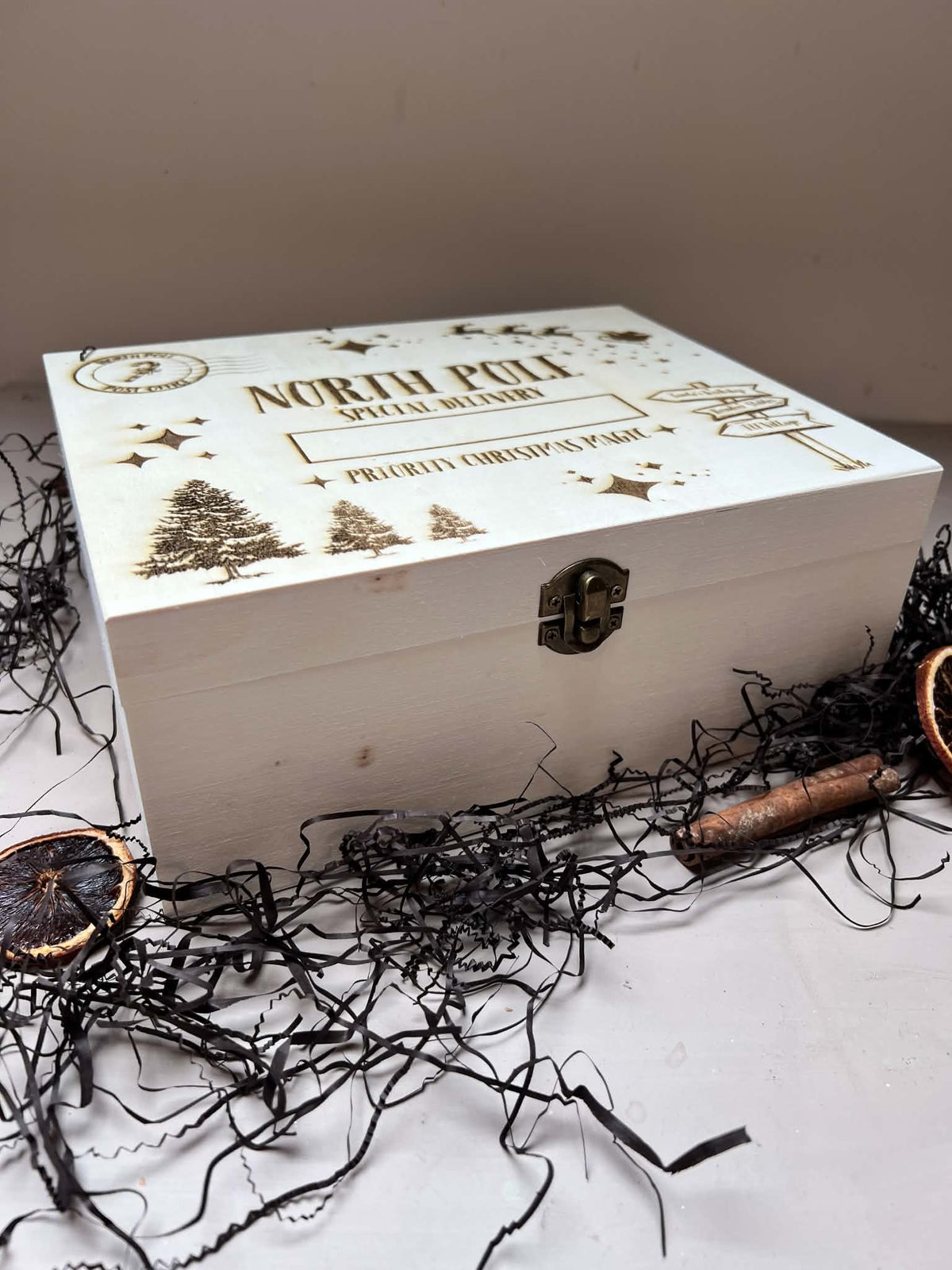 A white wooden gift box with a metal clasp lid, engraved with 'NORTH POLE SPECIAL DELIVERY' and Christmas tree illustrations, styled on a white surface with black shredded paper and dried orange slices.