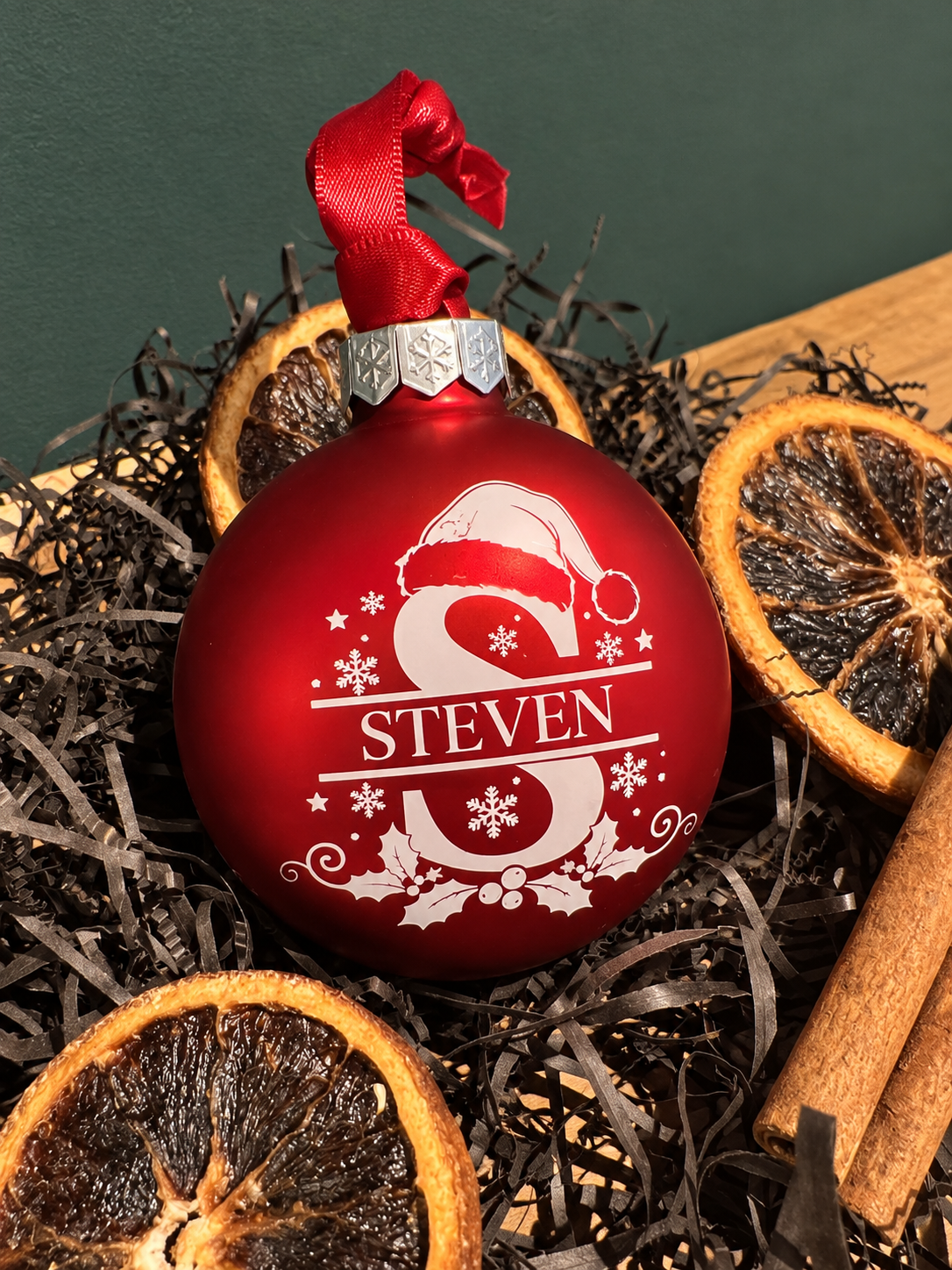 A red Christmas ornament with a silver top cap and red hanging ribbon, decorated with a large white monogram letter S, a Santa hat, snowflakes, and the name STEVEN. The ornament is styled in dark shredded paper with dried orange slices and cinnamon sticks against a dark green background.
