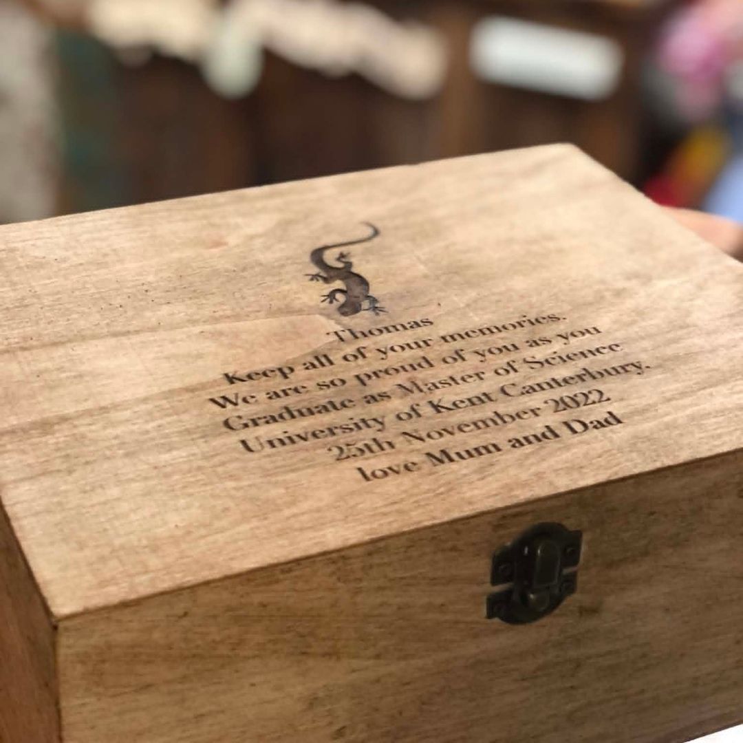 A close-up of a personalized wooden keepsake box with a small black metal latch on the front. The lid is engraved with a black ginkgo leaf symbol and a custom message for 'Thomas' from 'Mum and Dad' referencing graduation from Kent Canterbury University in 2022.