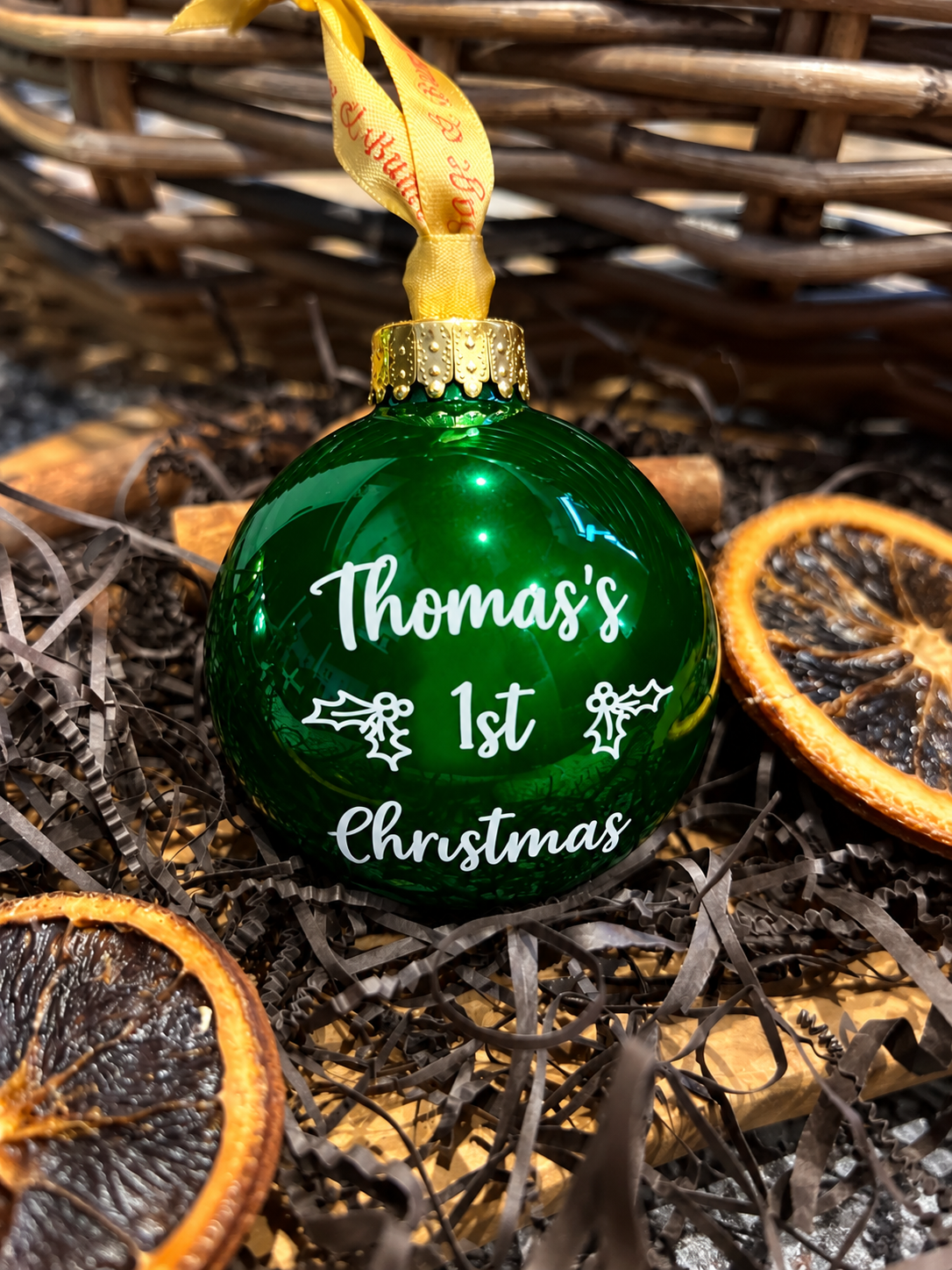A green glass Christmas ornament with white text reading 'Thomas's 1st Christmas' and a gold hanging ribbon, styled in dried orange slices and brown shreds near a wicker basket.