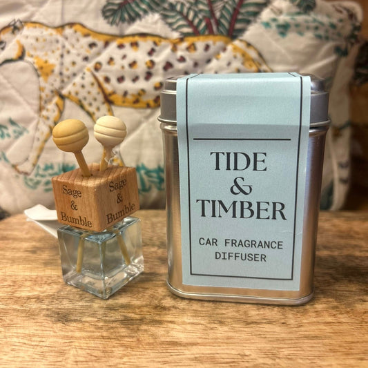 A Sage and Bumble Tide and Timber handcrafted car air freshener.  The car air vent diffuser bottle shown next to the metal tin on a wooden table.