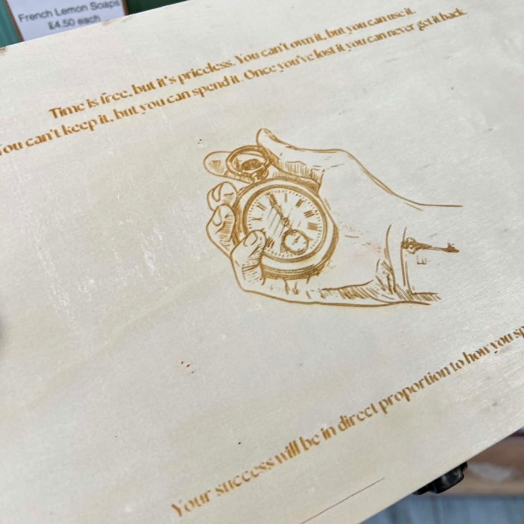 A close-up of a light wood box or plaque featuring a laser-engraved illustration of a hand holding a pocket watch, with motivational text printed on the surface.