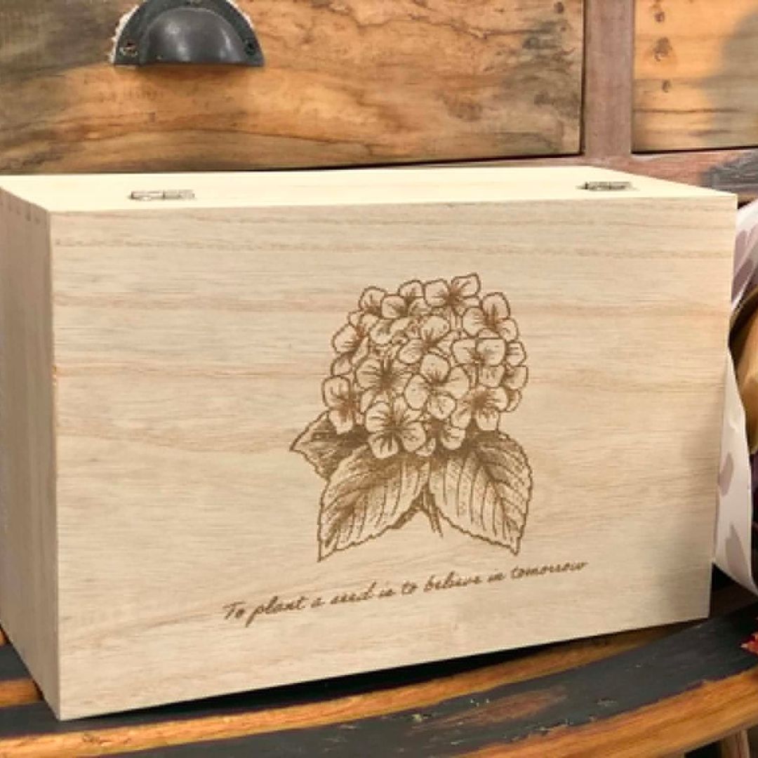 A rectangular light wood box with a laser-engraved illustration of a hydrangea and the quote 'To plant a seed is to believe in tomorrow' on the front, set against a rustic wooden background.