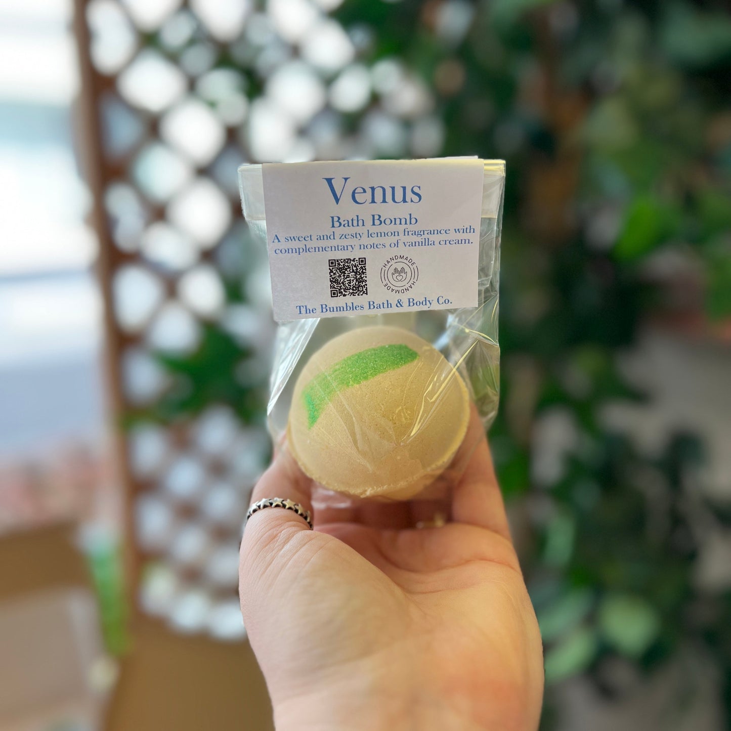 A hand holding a small clear plastic bag containing a round bath bomb labeled 'Venus Bath Bomb' by The Bumble, Bath & Body Co., with a blurred outdoor background of greenery and a lattice screen.